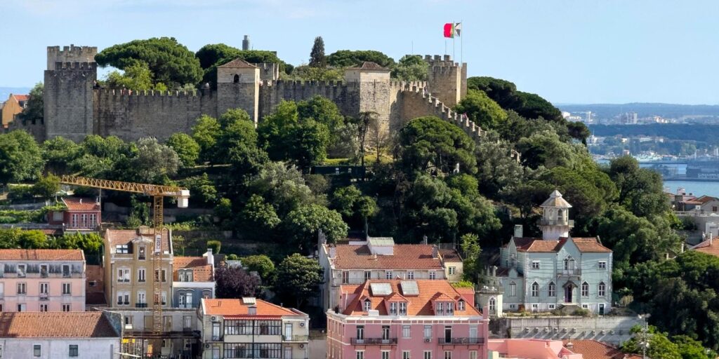 São Jorge Castle rising above Lisbon’s Alfama district with the Portuguese flag flying, surrounded by colorful houses and greenery — a must-see in this Lisbon travel guide.