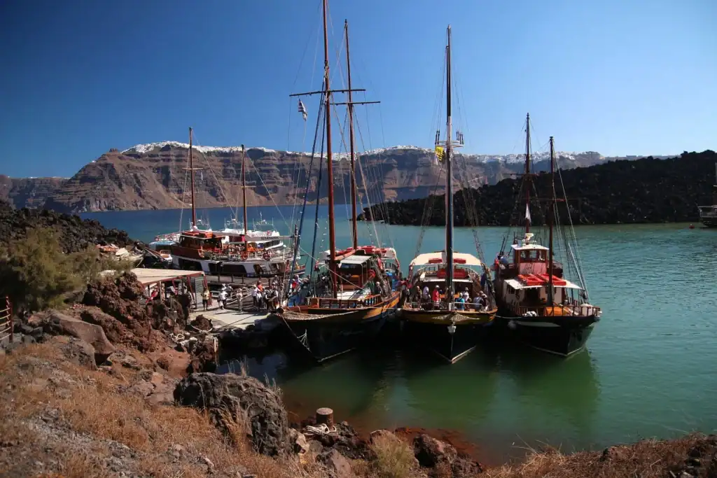 Santorini Travel Guide Nea Kameni volcanic islet seen from Santorini’s caldera, with moored boats and steep cliffs in the background.