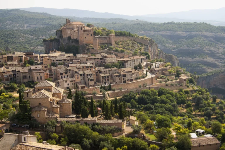 Wide landscape view of the medieval stone village of Alquézar in Aragon, Spain, with its hilltop fortress overlooking deep limestone canyon walls.
