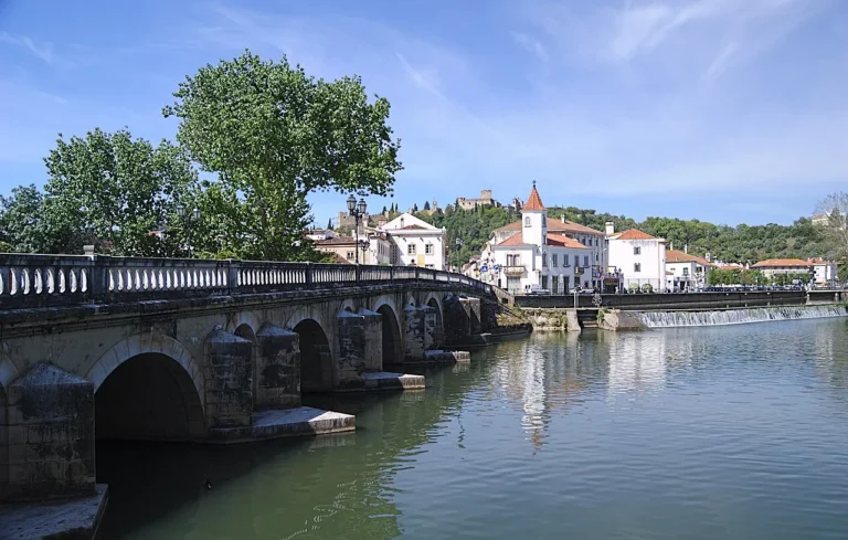 Historic arched stone bridge crossing the Nabão River in Tomar Portugal with the medieval Templar castle visible on the hill in the background.