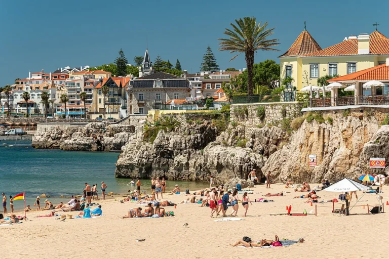 A vibrant, sunny view of Praia da Rainha beach cove in Cascais, Portugal, showing sunbathers on the golden sand, clear calm water, and historic grand villas built into the rocky seaside cliffs.