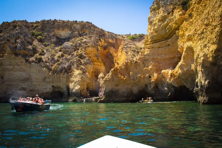 A boat navigating the clear emerald waters next to the golden limestone sea cliffs and cave entrances in Lagos, Algarve, Portugal.