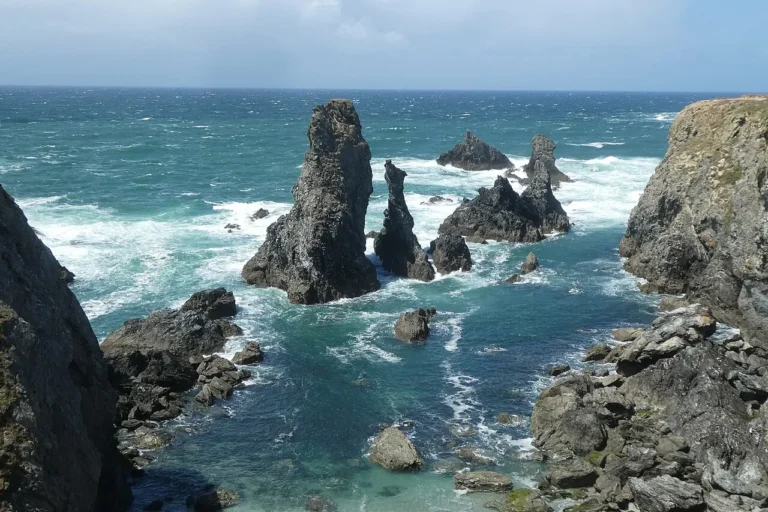 Dramatic, jagged granite rock stacks piercing through churning white ocean waves along the rugged, windswept coastline of Belle-Île-en-Mer, France.