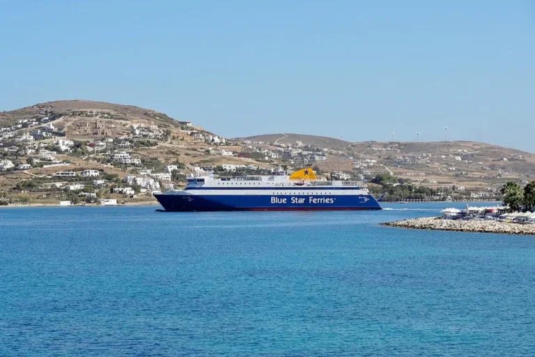 A large blue and white Blue Star Ferries passenger ship sailing into the turquoise waters of Parikia Bay on the island of Paros, Greece.