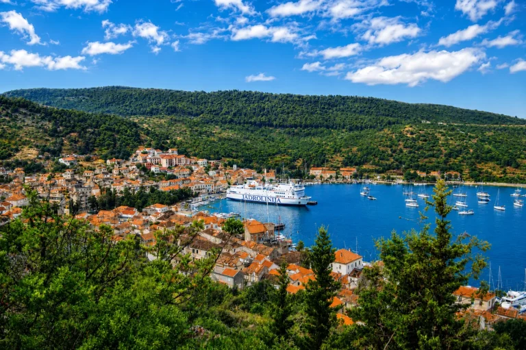 Scenic elevated view of a large car ferry docked at the harbor pier in Vis Town, Croatia, surrounded by terracotta roofs, sailing boats, and lush green hills.